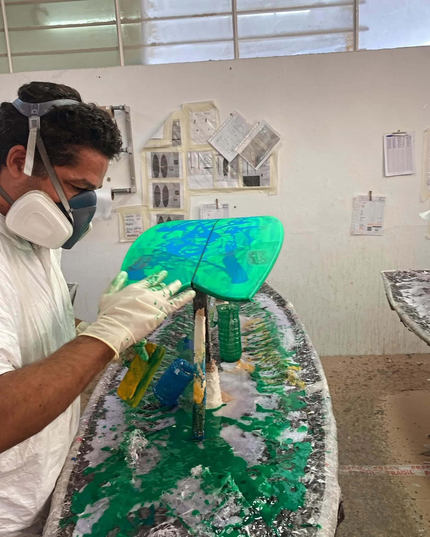 The builder in a respirator and suit, hand-laminating green tinted resin over a twin-fin in the glassing bay