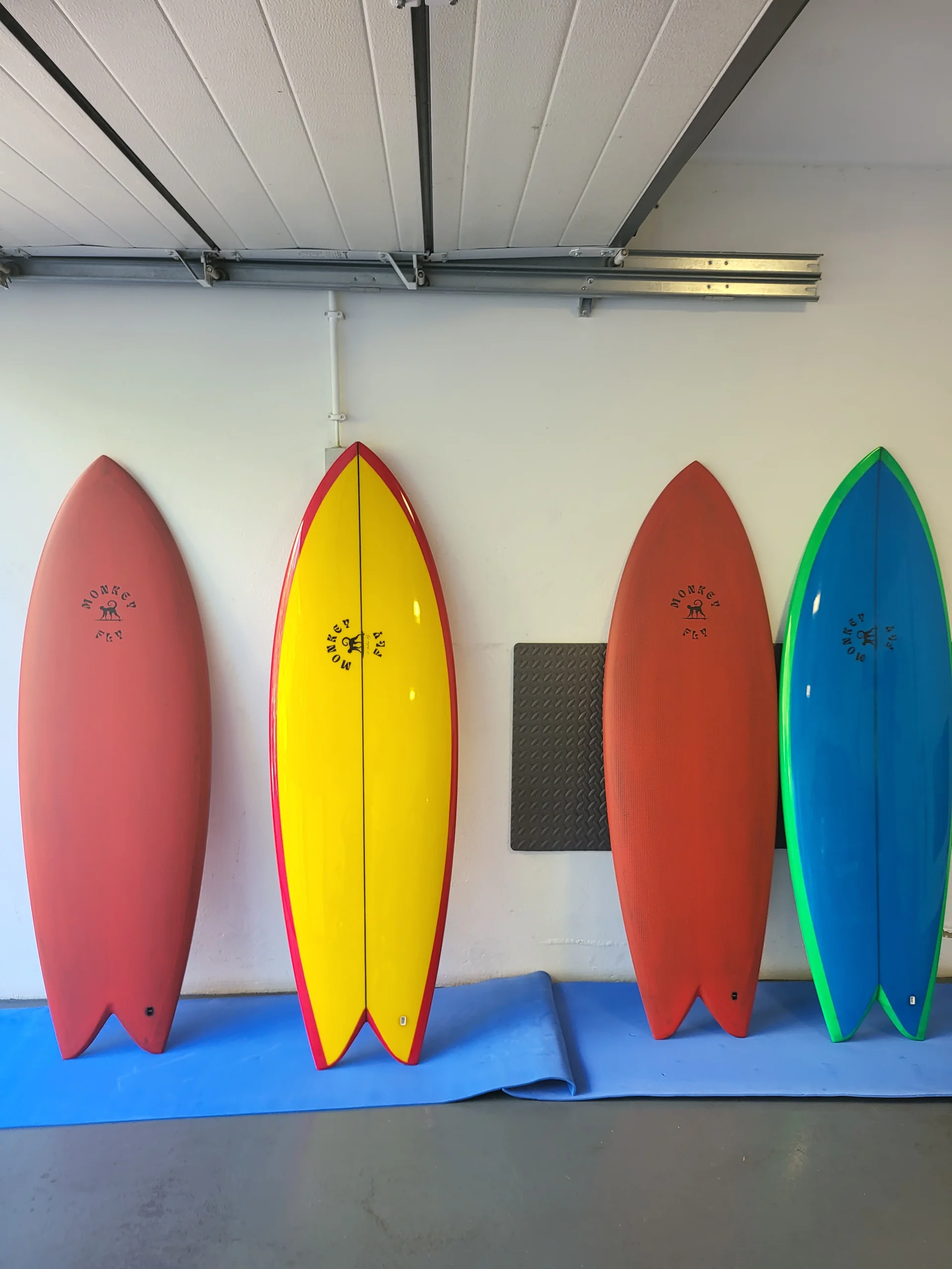 Four custom twin-fin surfboards in red, yellow, rust and blue resin tints standing on a workshop rack